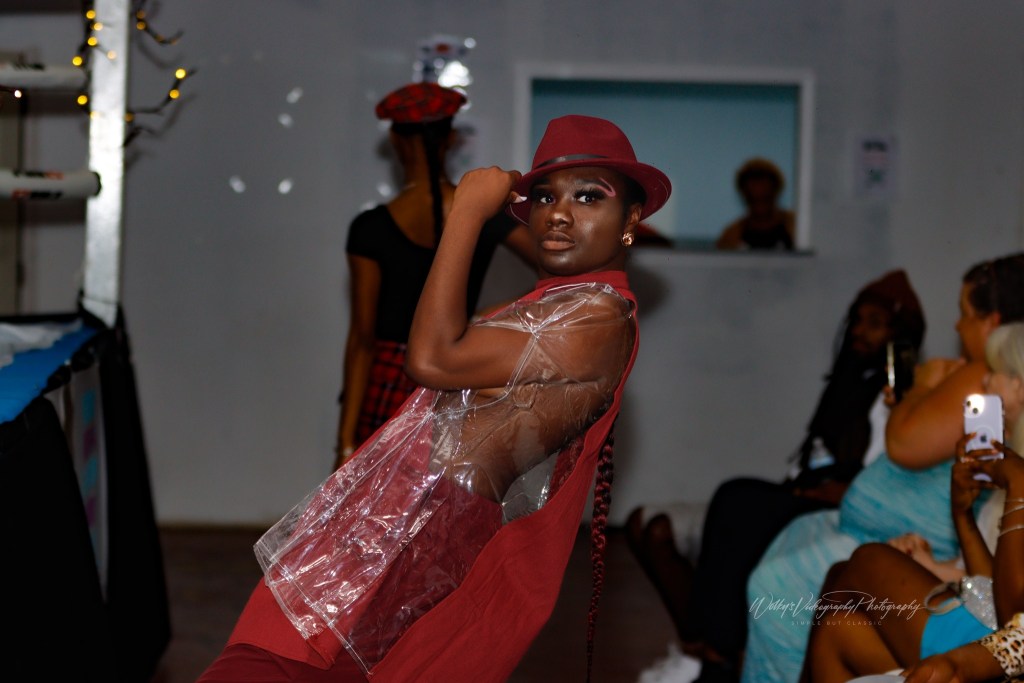 A model showcases a unique outfit, featuring a transparent layer and a stylish hat, on a runway. The audience is visible in the background, engaged with the presentation.